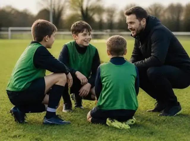 A youth football coach crouching to talk with young players on a grass pitch